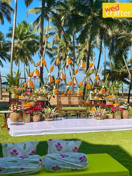 A vibrant Haldi ceremony setup on a sunny lawn by the beach, featuring a geometric bamboo backdrop decorated with marigolds and surrounded by colorful flowers.