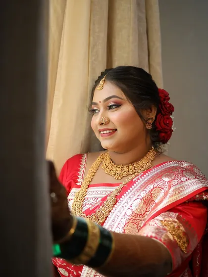 A happy bride looking out a window, her traditional red saree and gold jewelry catching the light beautifully.