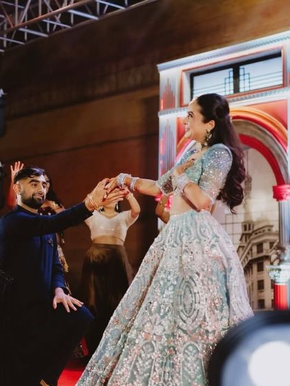 A kneeling pose that adds a touch of drama and romance to the couple's sangeet performance. The stage lighting and backdrop enhance the cinematic feel of the moment.