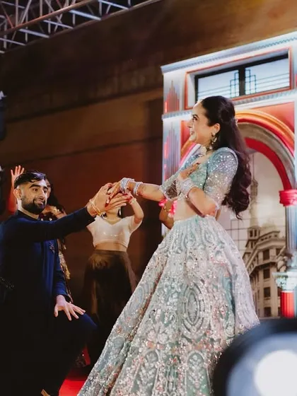 A kneeling pose that adds a touch of drama and romance to the couple's sangeet performance. The stage lighting and backdrop enhance the cinematic feel of the moment.