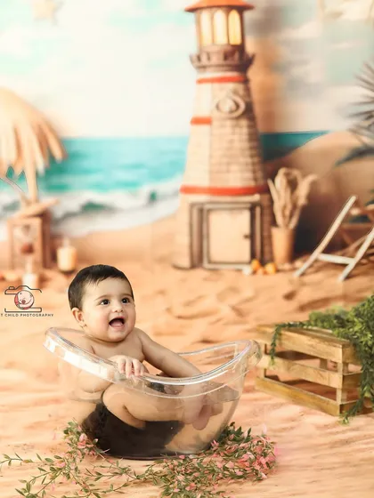 A happy baby enjoying a bath in front of a lighthouse. The detailed beach backdrop, complete with sand and greenery, makes for a very imaginative portrait.