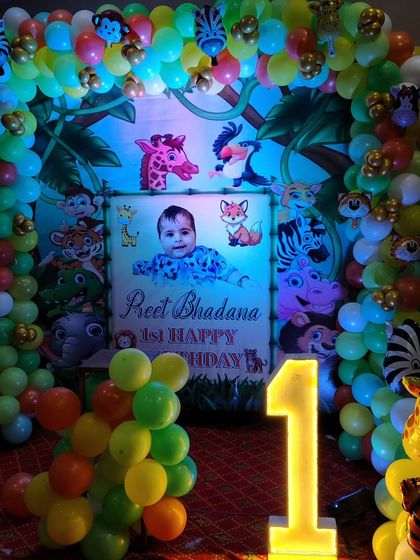 A closer look at a jungle-themed first birthday party. The setup includes a custom backdrop with the baby's name, a full balloon arch with animal prints, and a light-up number '1'.