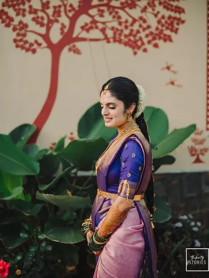 A serene bridal portrait against a backdrop of traditional wall art. The bride's calm demeanor and the beautiful contrast of her purple and pink saree create a stunning image.