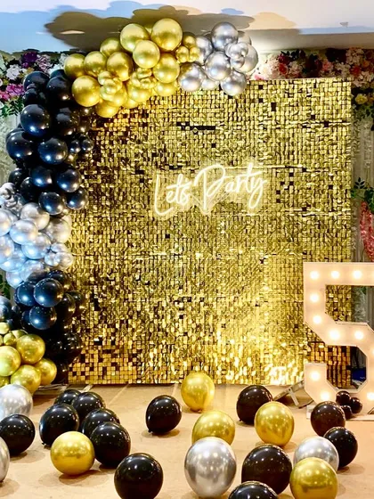 A chic and modern party setup for a 50th celebration. The decor features a bold black, gold, and silver balloon arch against a gold sequin backdrop with a "Let's Party" neon sign.