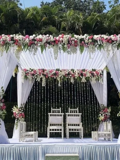 An elegant and classic outdoor wedding mandap design. This setup features a canopy of white drapes, lush pink and white floral arrangements, and delicate crystal hangings, creating a serene and beautiful space for the wedding ceremony.