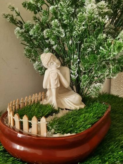 A beautiful Zen garden arrangement in a boat-shaped ceramic planter. It features a resting Buddha statue under an artificial tree, with a mini picket fence adding a touch of whimsy.