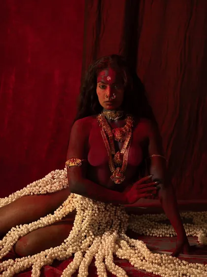 A full-body portrait from the 'Kali' series, showing the contrast between the red-painted skin and the white floral skirt. The pose is one of contemplative power.