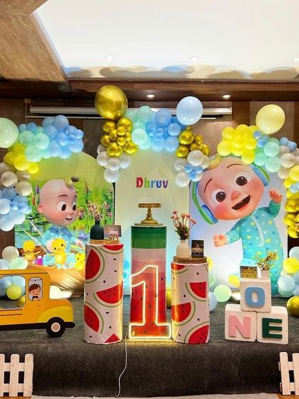 A Cocomelon-themed first birthday party. This elaborate stage setup includes character cutouts, a school bus prop, and a colorful balloon arrangement.
