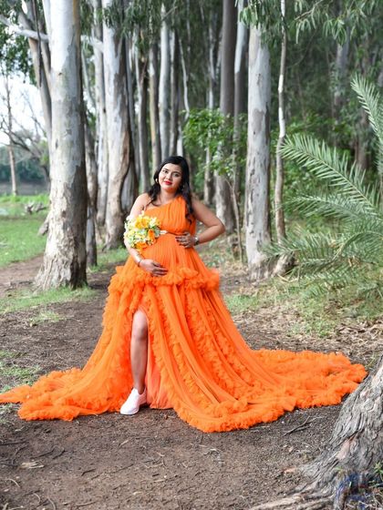 A bold and beautiful outdoor portrait. The mom-to-be is wearing a vibrant orange gown and white sneakers, holding a bouquet of yellow flowers in a forest setting.