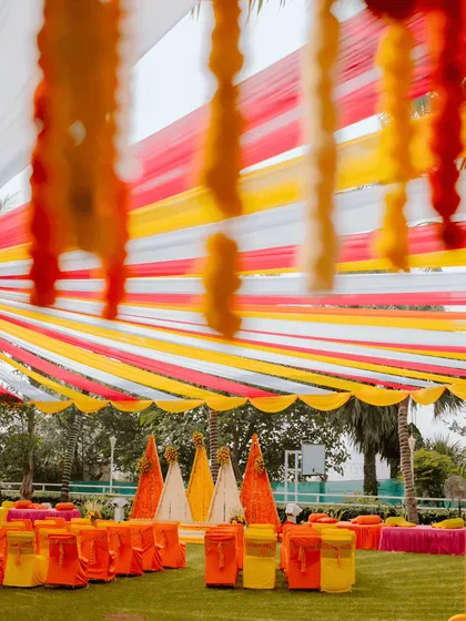 A wide view of a vibrant Haldi ceremony we designed. The space is brought to life with a canopy of colourful drapes in yellow, red, and white, complemented by marigold flower strings and cone-shaped floral backdrops, creating a festive and energetic outdoor setting.