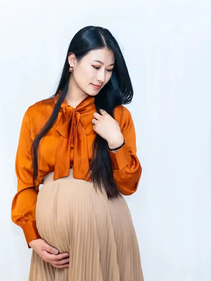 A graceful solo portrait of the mom-to-be. She looks down at her bump with a gentle smile, dressed in a stylish orange blouse and pleated skirt.
