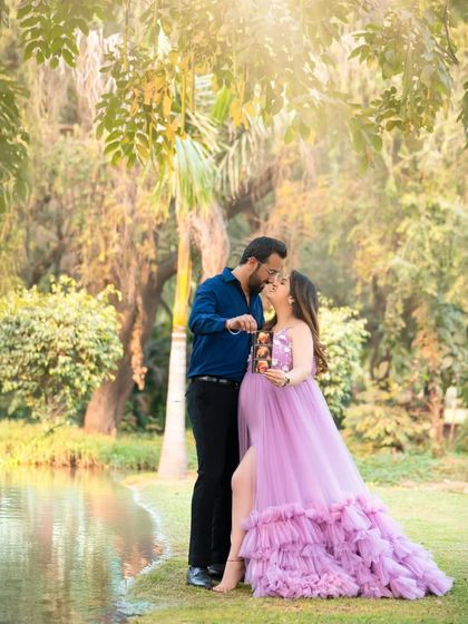A romantic outdoor portrait. The couple shares a kiss by the water, holding up their ultrasound photo in a beautiful, sun-drenched park.