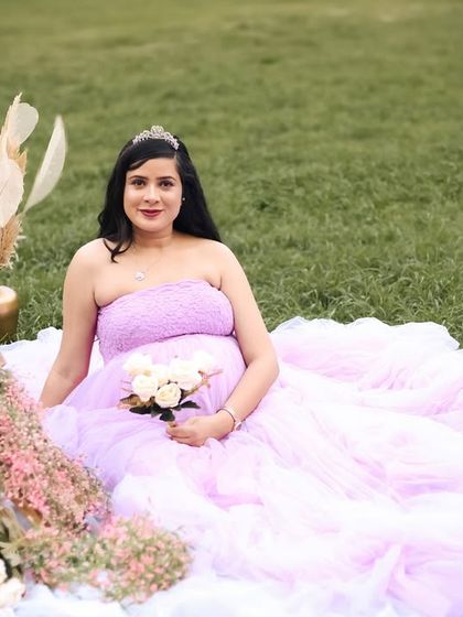 A lovely seated portrait from an outdoor session. The mother-to-be holds a bouquet of white roses, her lavender gown spread out on the grass, creating a romantic and serene scene.