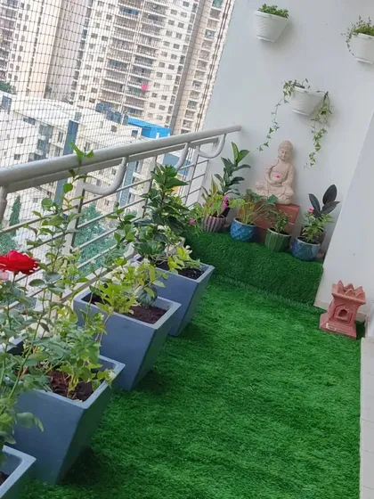This balcony garden design creates a serene corner with a mix of flowering plants in floor planters, wall-mounted pots, and a peaceful Buddha statue centerpiece, all set on a patch of artificial grass.