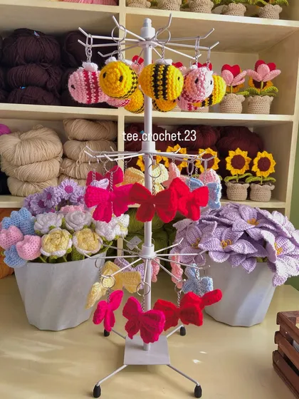 My display stand featuring bee and bow keychains, with colorful yarn and potted flowers in the background. This shows the range of cute accessories I create.