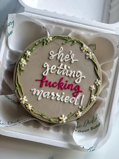 A cheeky and fun mini cake for a bride-to-be. The message "she's getting fucking married!" is framed by a delicate, hand-piped floral wreath.