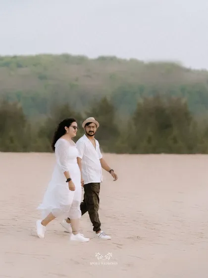 The couple taking a leisurely walk on a wide, sandy beach, with hills in the background.