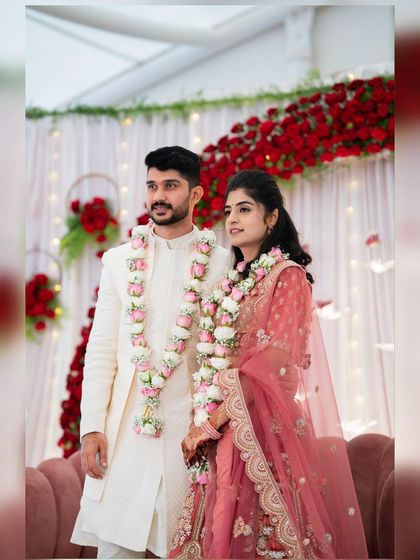 A lovely couple posing against the engagement stage we designed. The backdrop features a wall of red roses, white curtains, and delicate fairy lights, creating a warm and romantic ambiance for their ring ceremony.