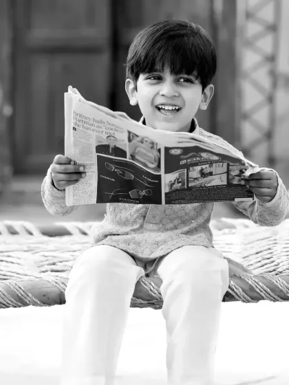 A happy moment captured in black and white, as a young boy reads a newspaper on a traditional charpai.