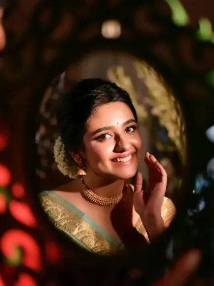 A creative shot using a mirror to frame the model's smiling face. Dressed in a beautiful green and gold saree, this image feels personal and joyful, capturing a moment of self-adornment.