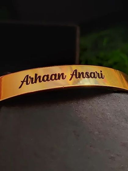 A close-up of a gold-plated cuff engraved with 'Arhaan Ansari'. The bracelet is resting on its gift box, showing the premium packaging it arrives in.