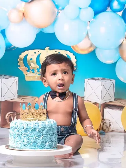 A little prince celebrating his first birthday with a royal blue cake smash. The setup includes a crown, balloons, and a delicious-looking cake ready to be smashed.