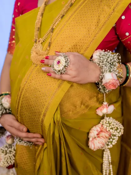 A close-up detail shot focusing on the mother-to-be's hands cradling her baby bump. This image highlights the beautiful textures of her golden saree and the intricate floral jewelry, emphasizing the precious details of her pregnancy.