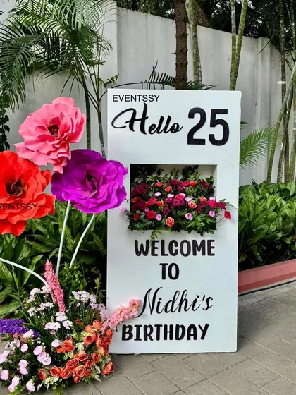 The personalized welcome sign for the 25th birthday party, saying "Hello 25." The sign features a floral shadow box and is accented with large paper flowers, creating a stylish first impression for guests.