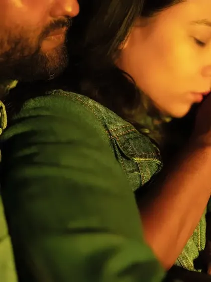 A close-up of the couple huddled together for warmth in the cool night air. The soft, yellow light from the campfire illuminates their faces, highlighting their closeness and affection.