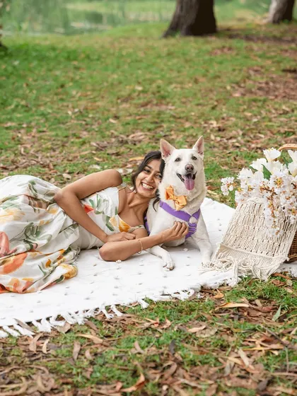 A joyful moment from an outdoor Mother's Day shoot. A woman lies on a picnic blanket with her happy white Indie dog, both smiling brightly.