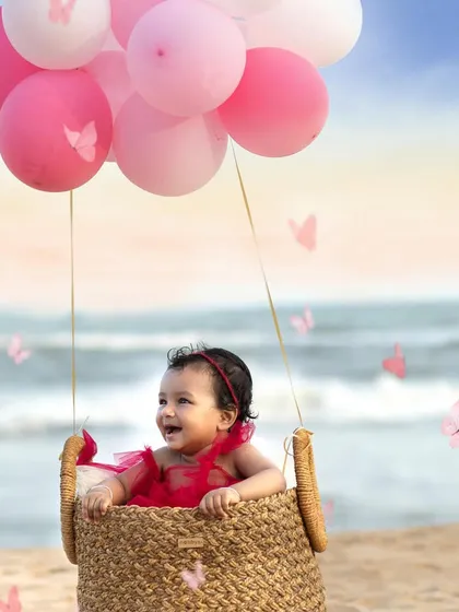 A whimsical and creative beach portrait of a baby girl smiling in a basket "hot air balloon" lifted by pink balloons.