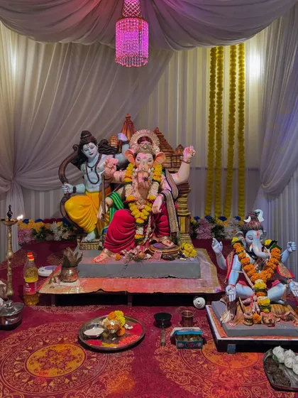 A full view of the pandal setup for the 'Gokul Nagari Cha Maharaja' 2024 event. This shows the simple yet clean arrangement with white fabric draping, a pink chandelier, and a well-lit space for multiple idols.