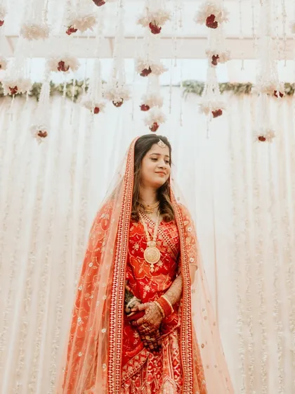A classic bridal portrait. The bride stands gracefully under the floral mandap, her red lehenga vibrant against the white backdrop. A picture of elegance and poise.
