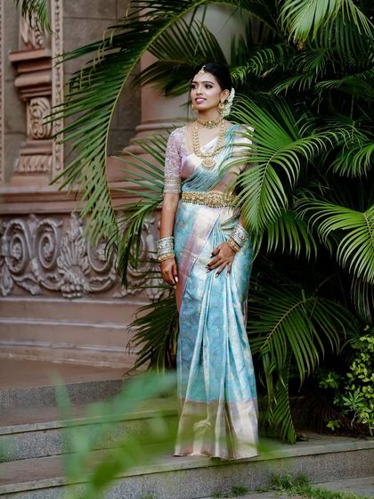 A bride stands gracefully amidst palm trees in a beautiful pastel blue and pink silk saree. The blouse is heavily embroidered, adding a touch of glamour to the soft-hued saree.