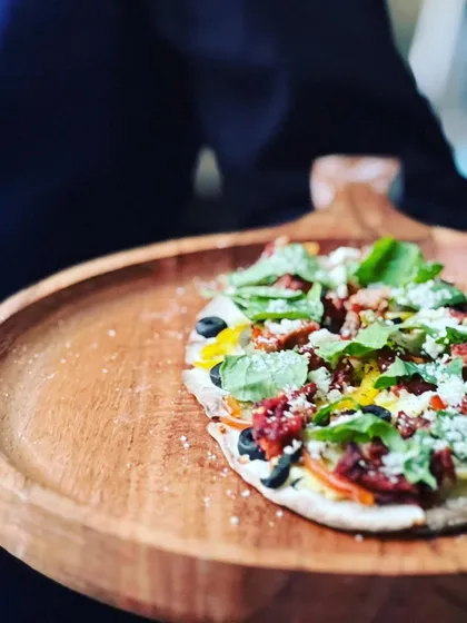 A freshly baked flatbread pizza, topped with sun-dried tomatoes, olives, and fresh basil, served on a rustic wooden board.