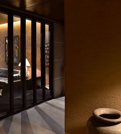 A glimpse into a living space through a screen of vertical wooden slats. The warm, low lighting creates an intimate and moody atmosphere, hinting at a comfortable room beyond.