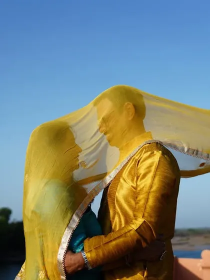 A romantic moment from an anniversary shoot, with the couple sharing a look under a flowing yellow dupatta against a clear blue sky.
