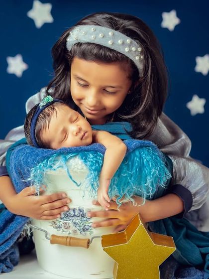 A big sister lovingly holds her sleeping newborn sibling against a starry blue backdrop. A magical and heartwarming newborn and sibling portrait.