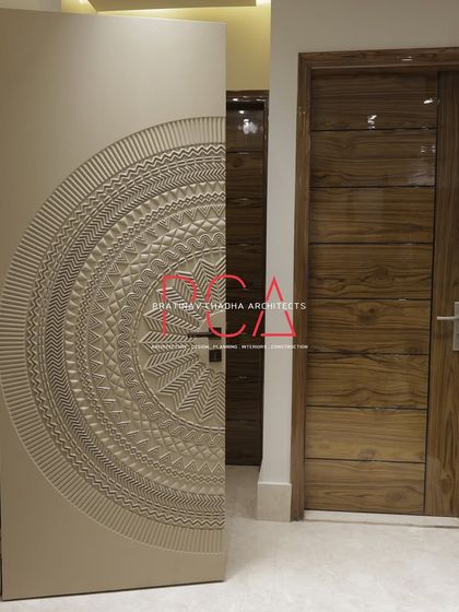 The bespoke main door of 'The Cursive Abode', shown here slightly ajar. The intricate exterior carving contrasts with the clean, modern wooden door of the adjacent room, highlighting a blend of styles.