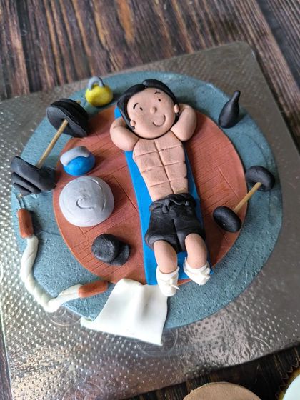 A gym-themed cake for a fitness lover. This design features a fondant figurine showing off his abs, surrounded by tiny, handcrafted gym equipment like dumbbells and kettlebells.