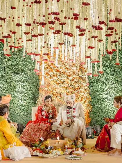 The family seated at the mandap, with the backdrop of green foliage and a checkered pattern of marigolds adding a traditional texture.