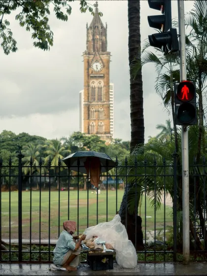 A street vendor sits by the roadside under an umbrella, with the Rajabai Clock Tower in the distance. The red pedestrian signal adds a pop of color and a sense of pause to the scene.