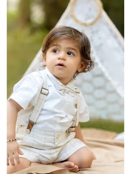 A classic portrait of a little boy during his pre-birthday twin photoshoot. Dressed in a smart shirt and suspenders, he sits in front of a lace teepee, looking thoughtfully into the distance.