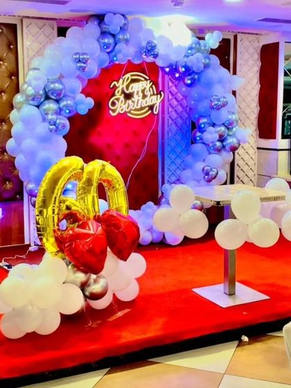 A grand setup for a 60th birthday celebration in a banquet hall. The decor features a silver balloon arch on a red stage, with large "60" foil balloons, creating a celebratory and honorable atmosphere.