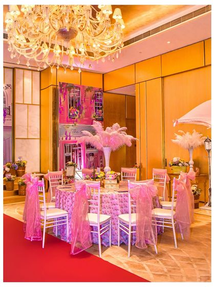 An elegant dining table at the Parisian party, with pink draped chairs, a textured tablecloth, and a large feather centrepiece, all set against the backdrop of the custom-designed Parisian street.