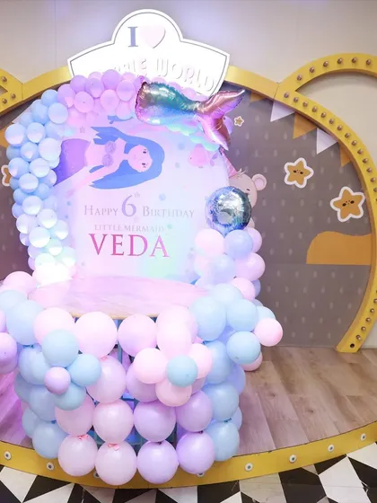 A beautiful mermaid-themed birthday setup. The decor features a shimmering mermaid tail, pastel balloon arrangements, and an underwater-inspired backdrop for "Little Mermaid Veda."