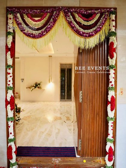 A beautiful main door decorated with multi-layered floral hangings ('thorana'). The rich colors and textures create a grand and festive welcome for a Gruha Pravesha.