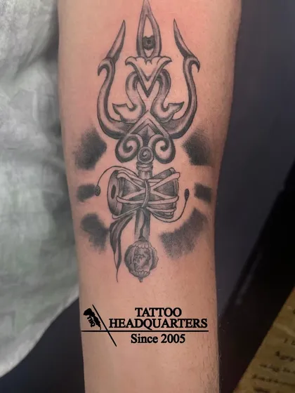 A black and grey tattoo of a Trishul with a Damaru, shaded to create depth and a powerful visual effect.
