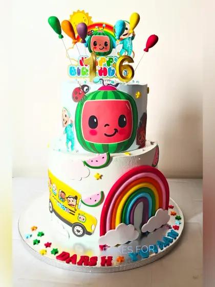 A vibrant two-tier Cocomelon cake, featuring JJ, the iconic watermelon, and the yellow school bus. The design is full of color with a big rainbow, perfect for a toddler's birthday.