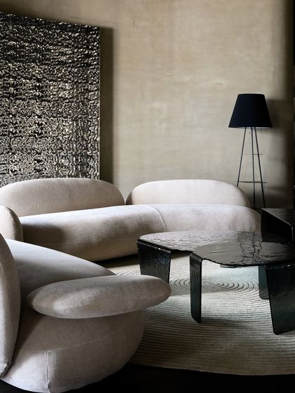 A minimalist living room setting featuring a low-profile sofa and armchair with soft, rounded forms. The textured wall art and dark, sculptural coffee table add layers of interest to the neutral palette.